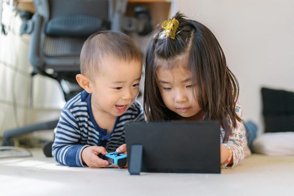 A small and cute baby boy is playing video games with his sister at home in the living room.