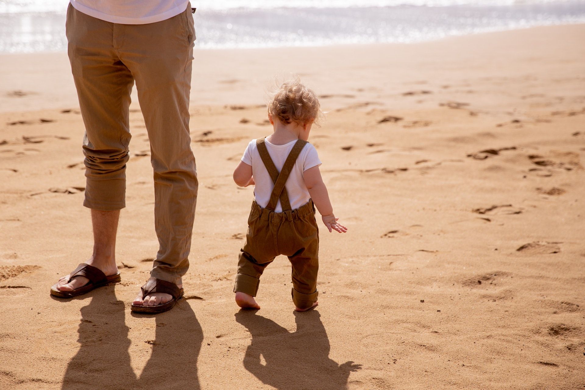 A toddler walks on the beach next to their father.